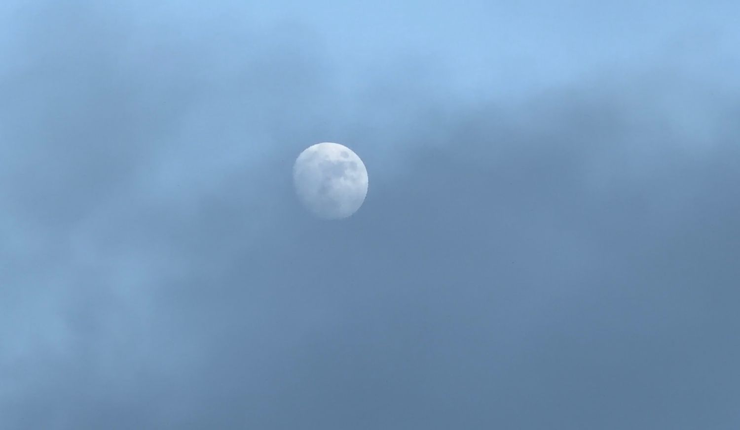A full moon glowing through soft blue-gray clouds in a twilight sky.