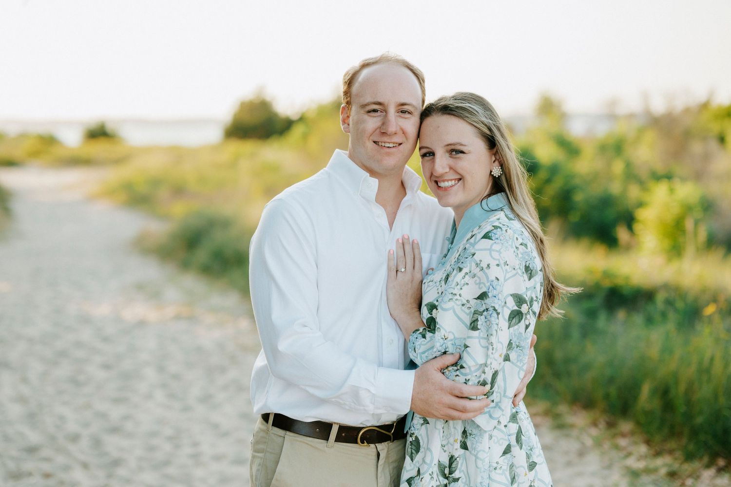 Couple smiling together on a sandy beach path surrounded by green foliage in warm golden light.
