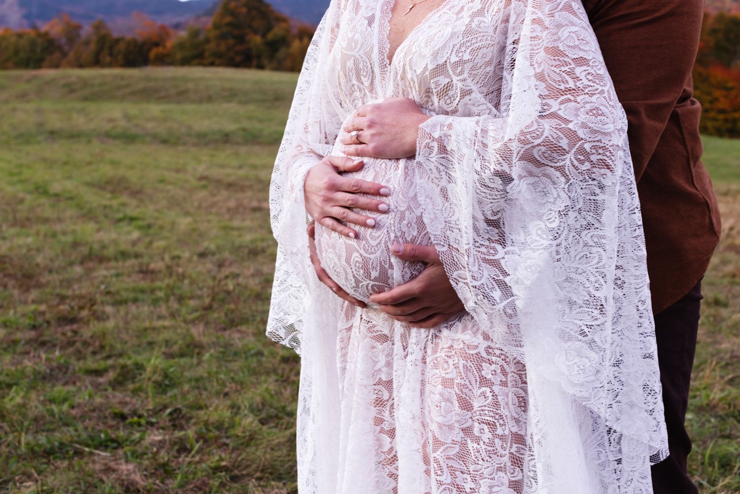 Expectant mother wears a white lace dress with fiancee embracing her from behind with an open field and fall foliage as a backdrop in Stowe.