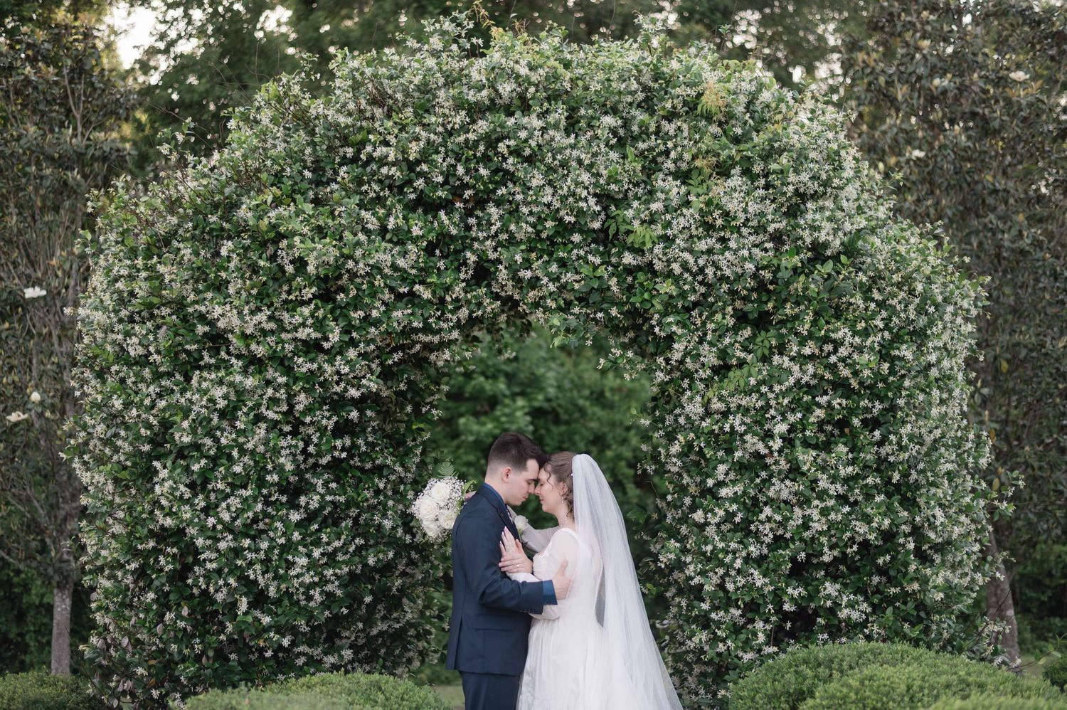 Bride and groom share romantic first dance dip on outdoor patio with string lights and lush green hedges backdrop.