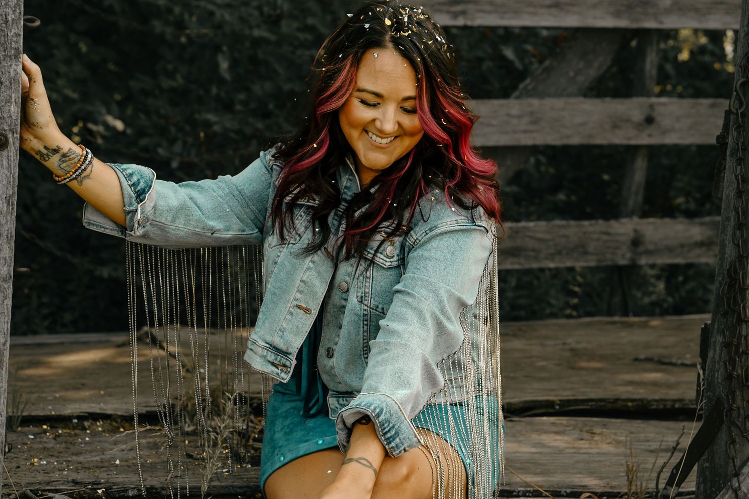 A person in denim jacket with colorful hair poses playfully on a wooden fence.