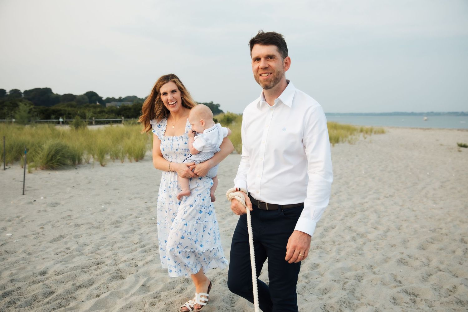 A couple enjoys a sunny day together walking along a sandy beach with grass and ocean in the background. , Middletown RI Third beach