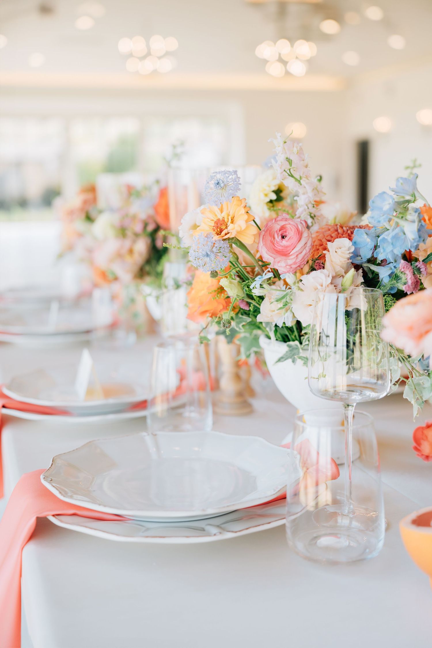 Elegant table setting with coral napkins and pastel floral centerpieces featuring pink and peach roses with blue accents.