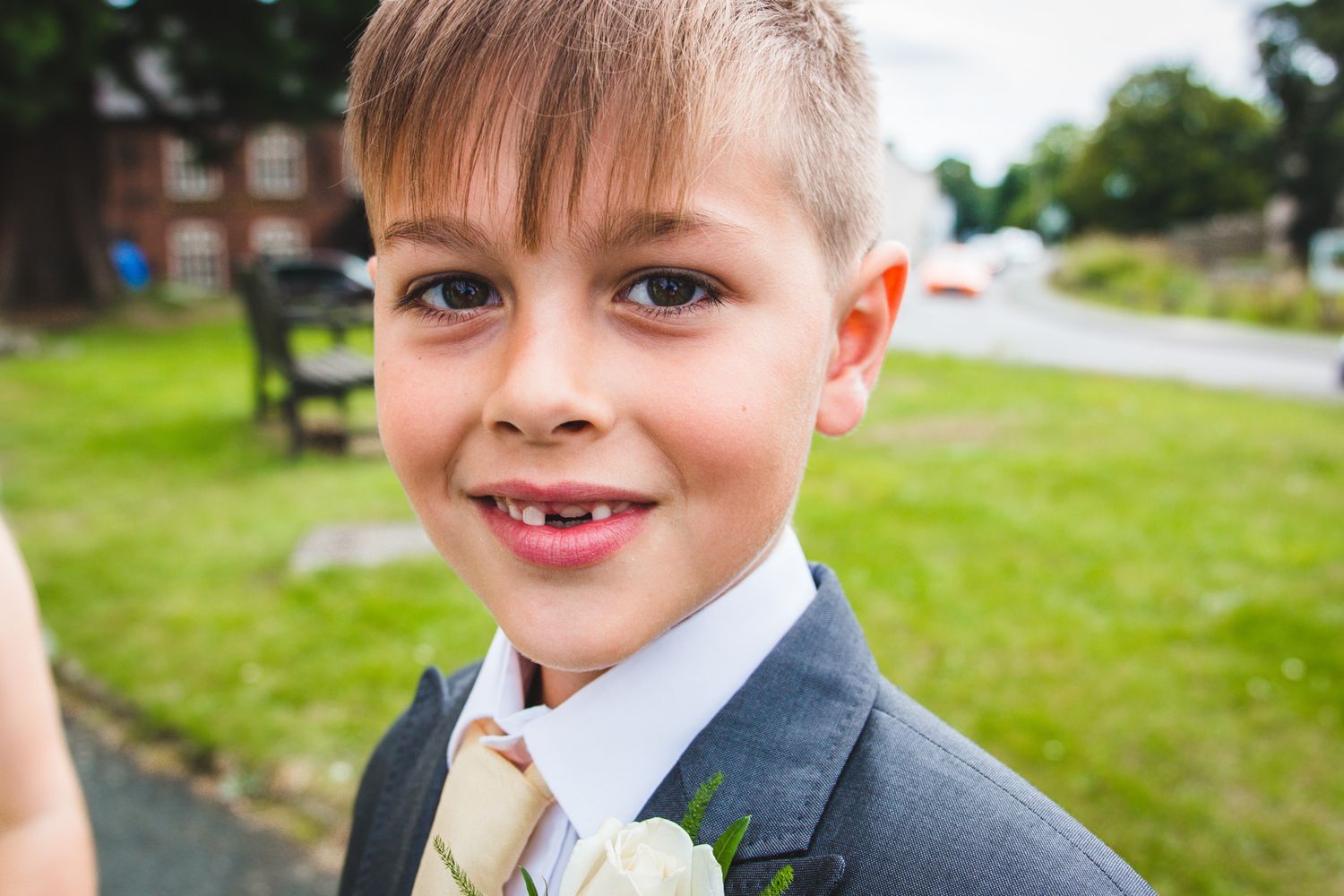 A young child wearing a navy suit and striped tie smiles brightly outdoors on a grassy lawn.