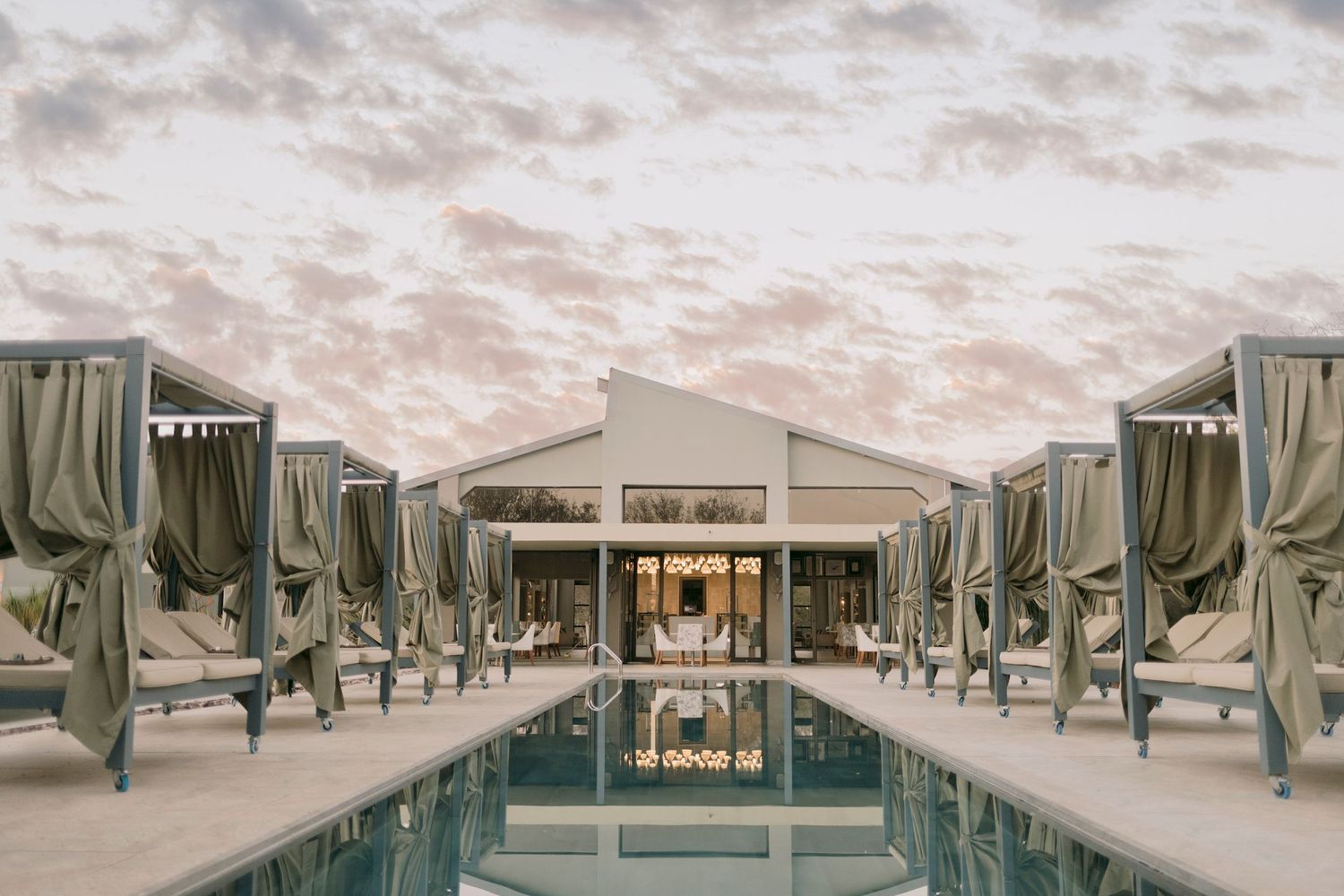 Luxury resort pool flanked by cabanas with sage curtains reflecting a pastel sunset sky.
