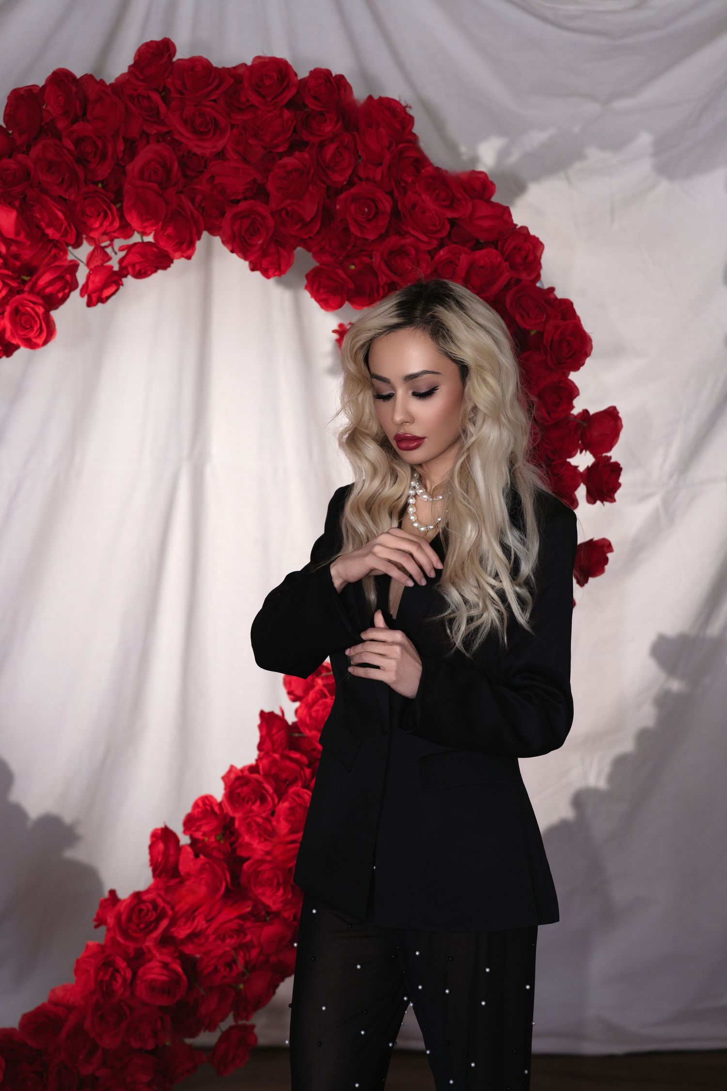 Fashion shoot featuring glamorous pose against dramatic red rose arch backdrop with white curtain and black attire.