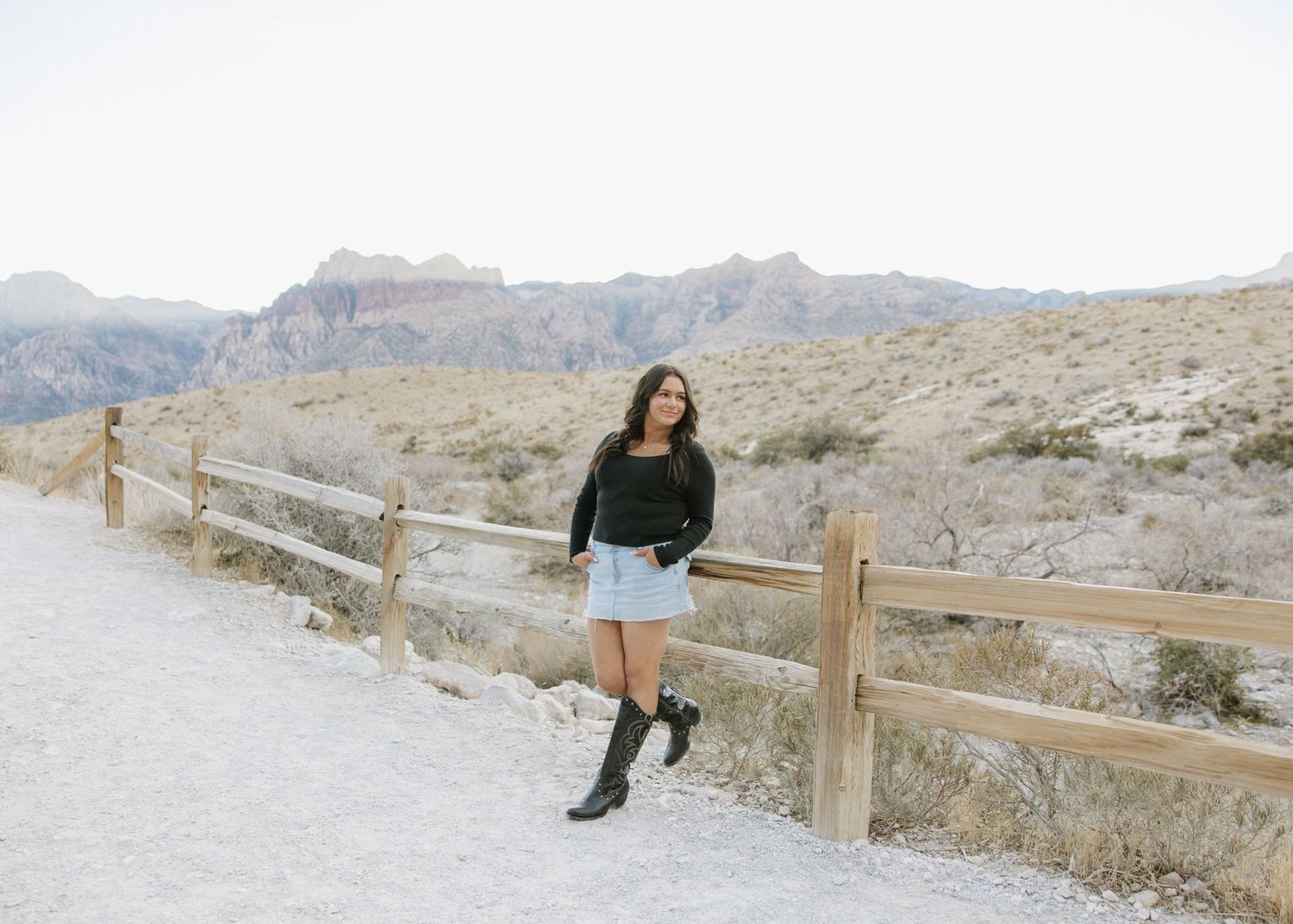 Person in black sweater and denim shorts stands by wooden fence with desert mountains in background.