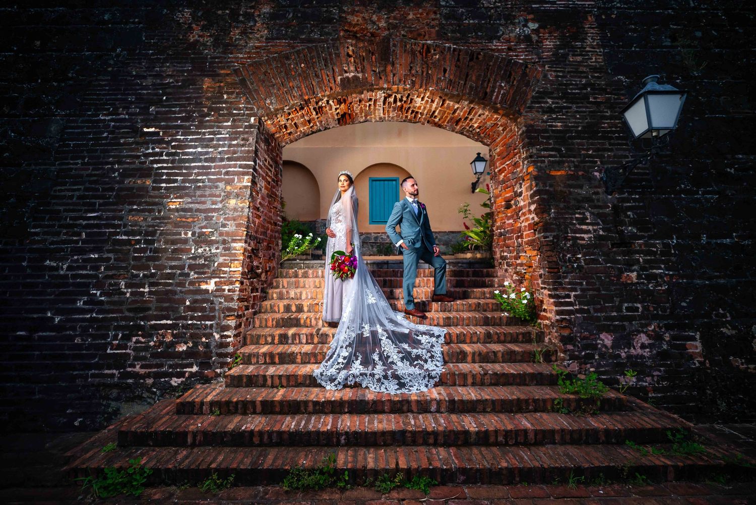 Wedding couple posing under dramatic brick archway with vintage lighting.