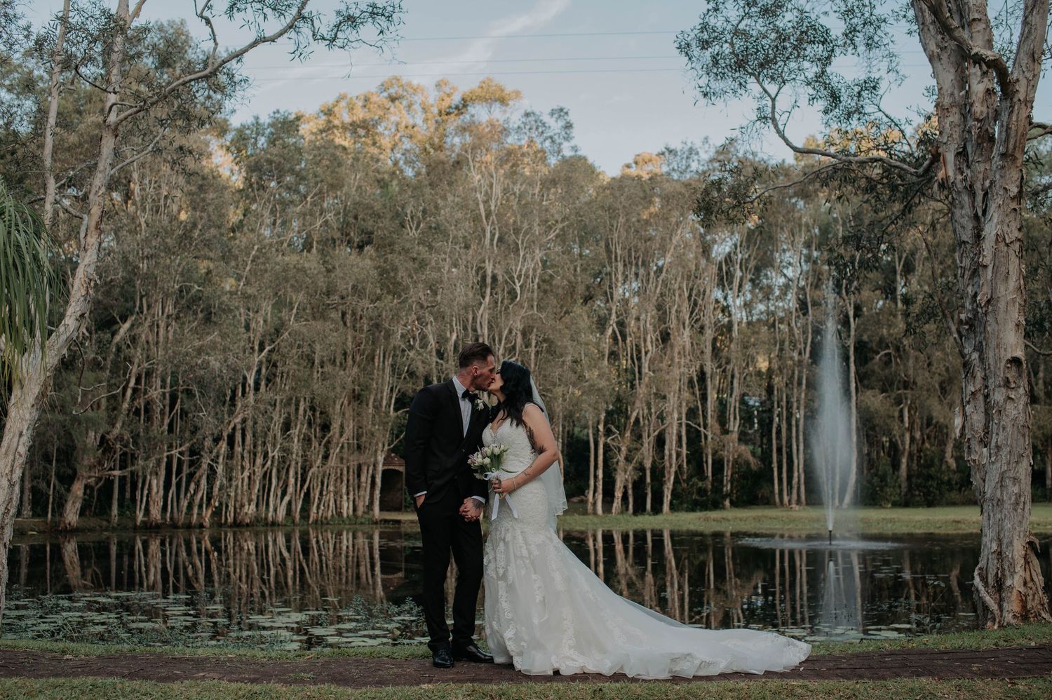 Two figures embrace in a romantic moment beside a serene lake surrounded by towering trees at sunset.
