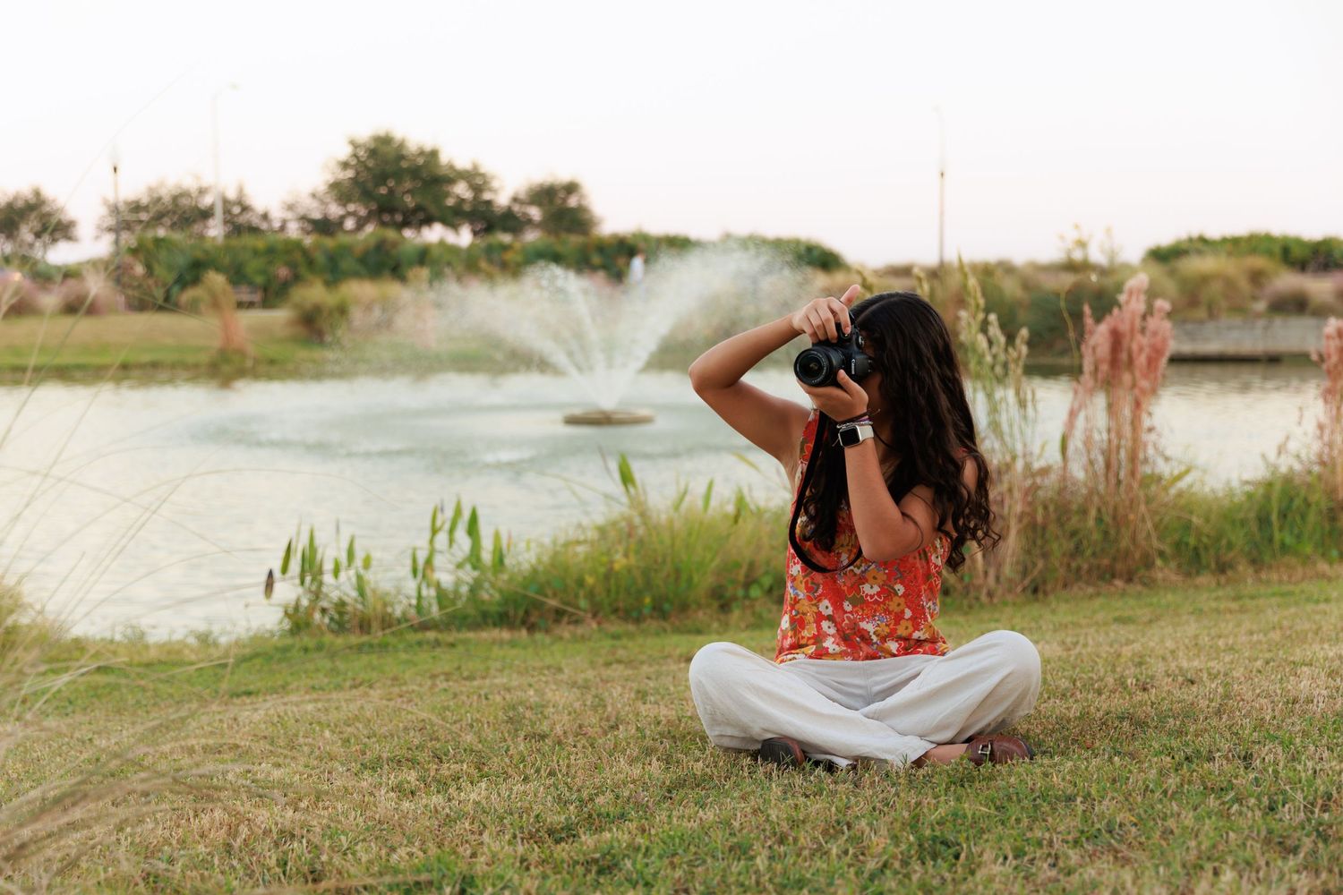 Photographer adjusts camera settings while sitting by pond with fountain spray in background.