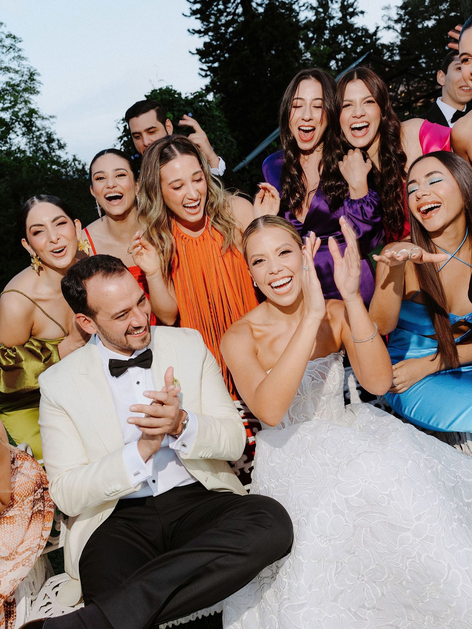A joyful wedding celebration with the newlyweds and wedding party laughing together outdoors.