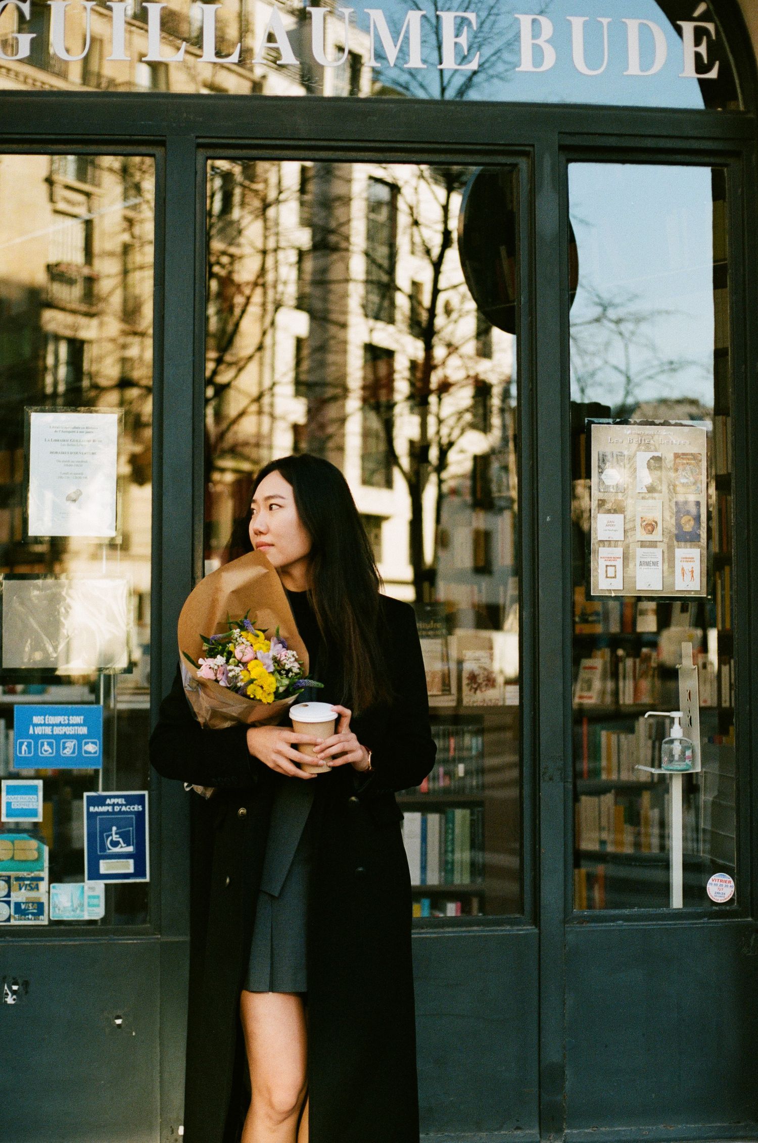 Woman in a black coat holding flowers and coffee stands outside a storefront with 'BUDE' signage in a Parisian setting.