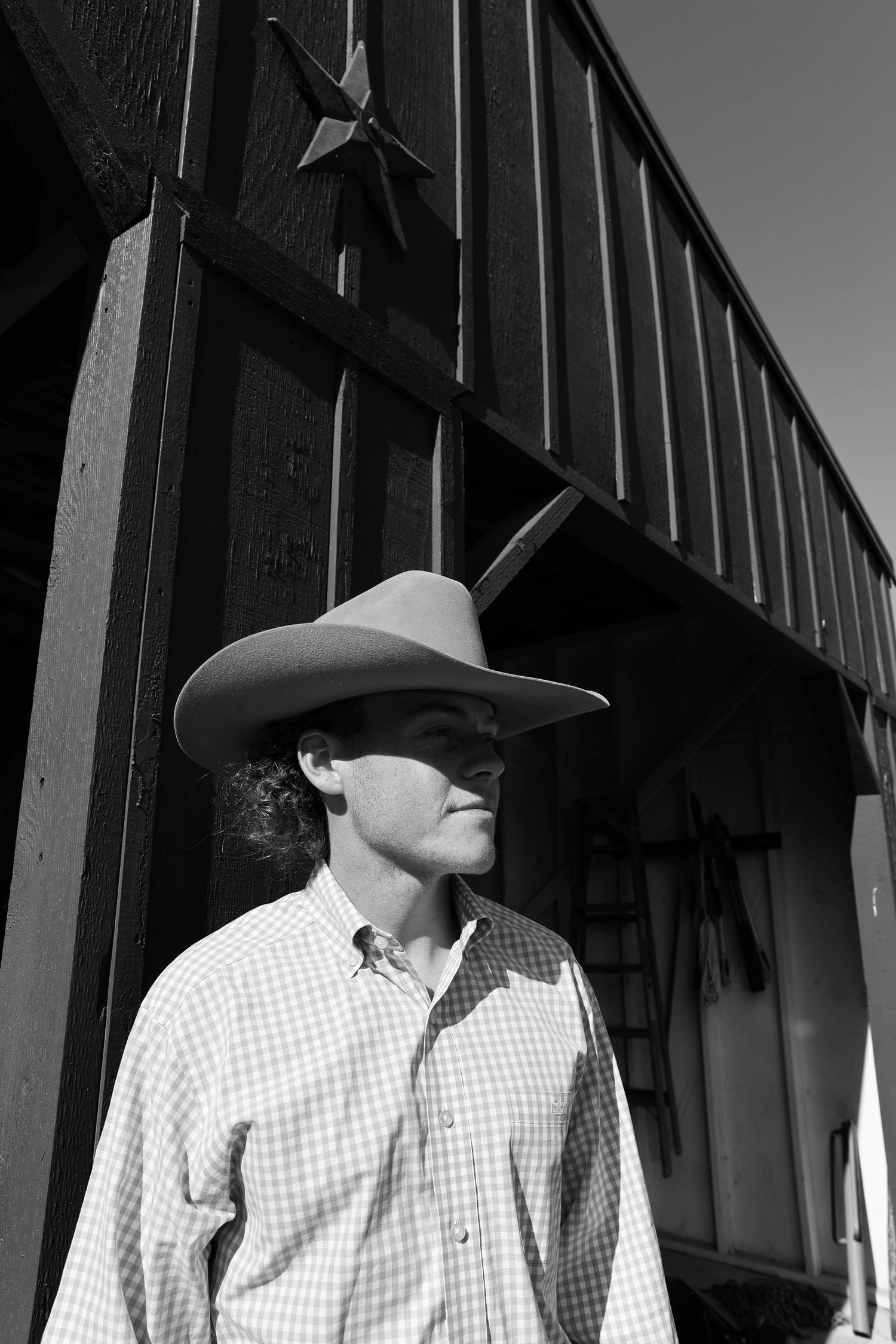 Black and white photo of someone in a cowboy hat and gingham shirt looking away while standing near a barn.