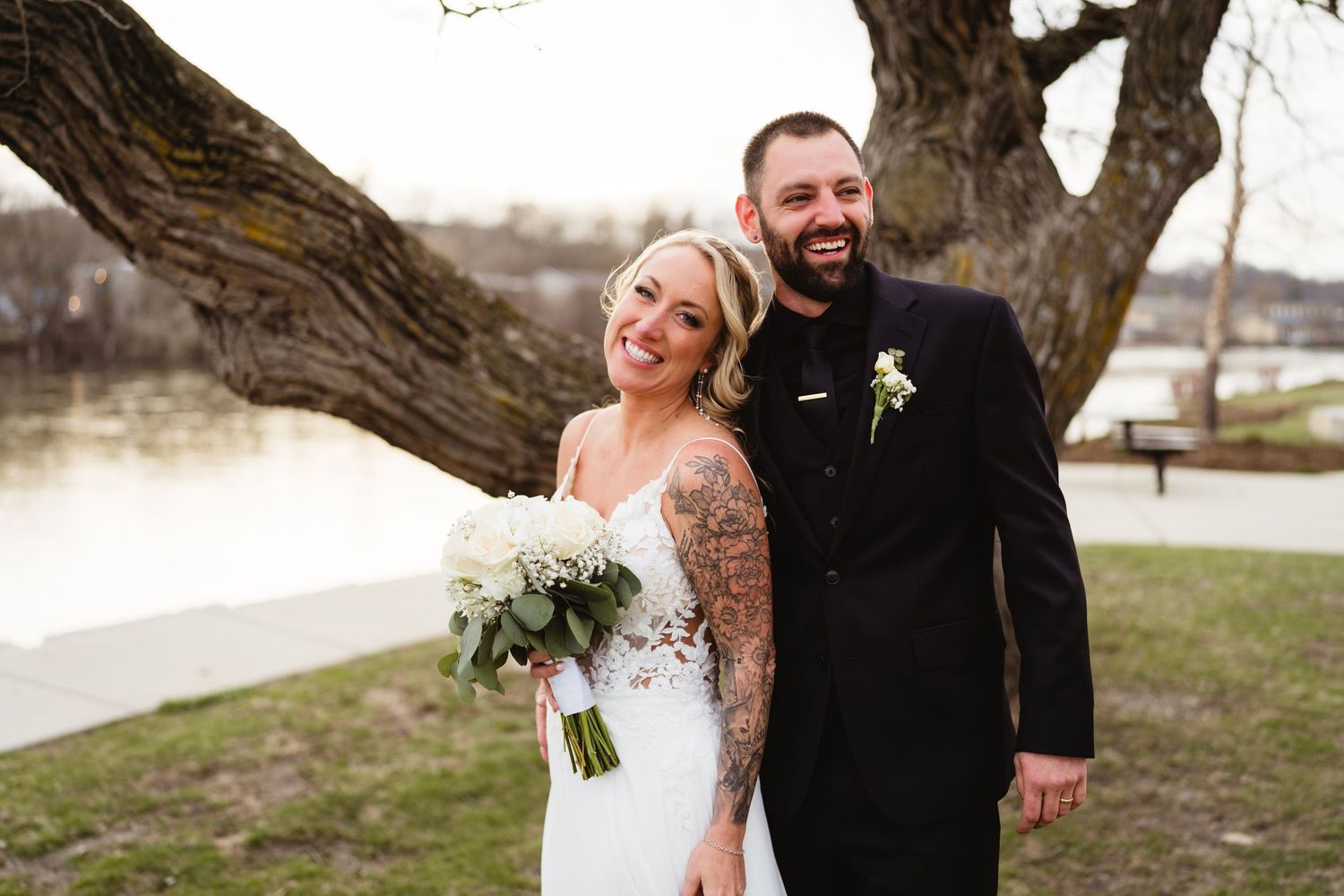 A happy newlywed couple embrace outdoors on their wedding day with the bride holding a white floral bouquet.