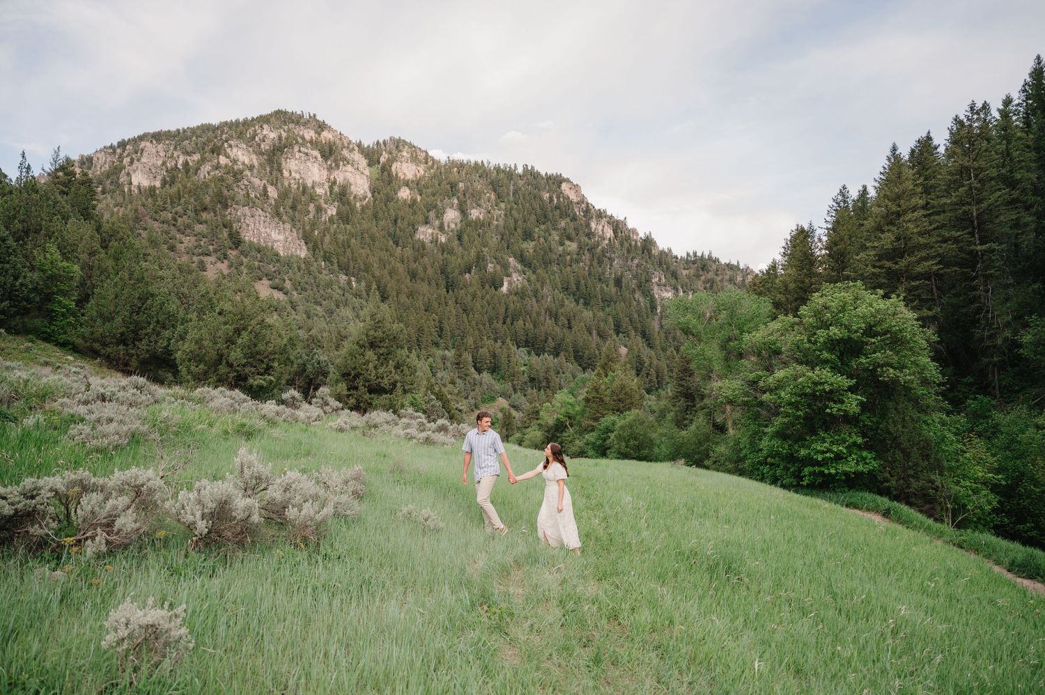 Two people walk hand in hand across a green mountain meadow with rocky cliffs and evergreen forest in the background.