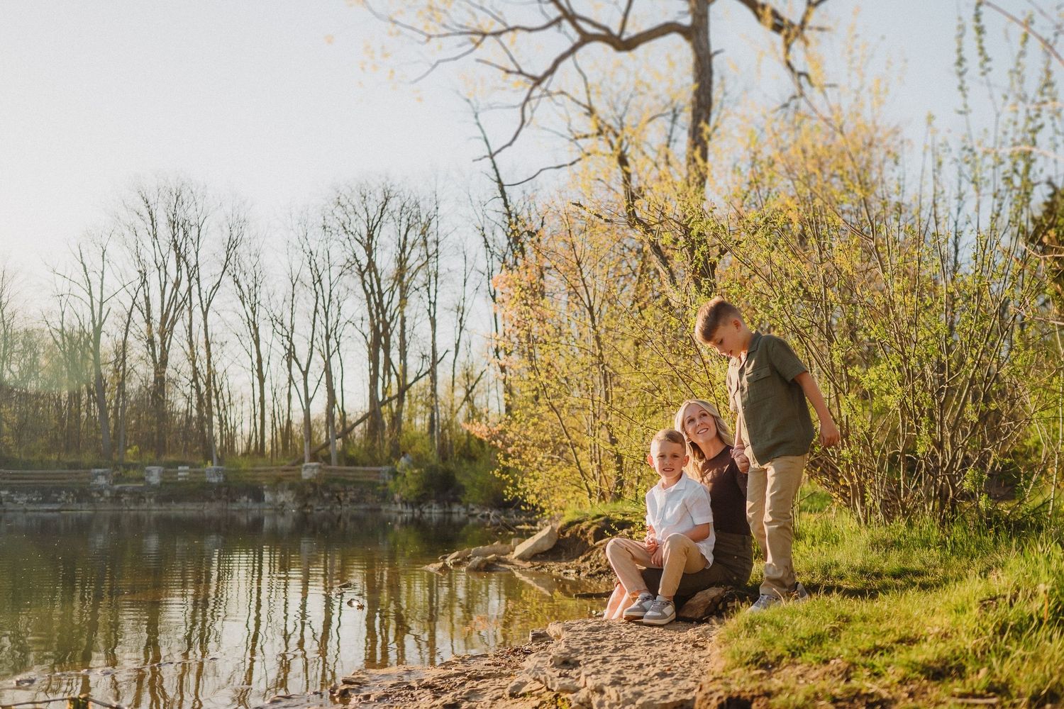 Two people sit together with a dog by a tranquil pond surrounded by autumn trees at sunset.