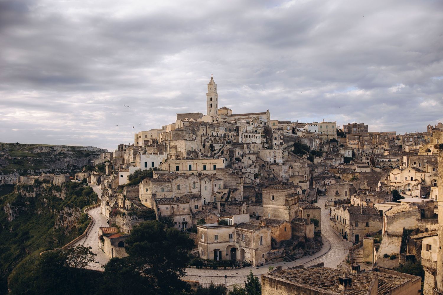 Ancient hillside town of Matera, Italy with stone buildings and cathedral rising dramatically against cloudy sky.