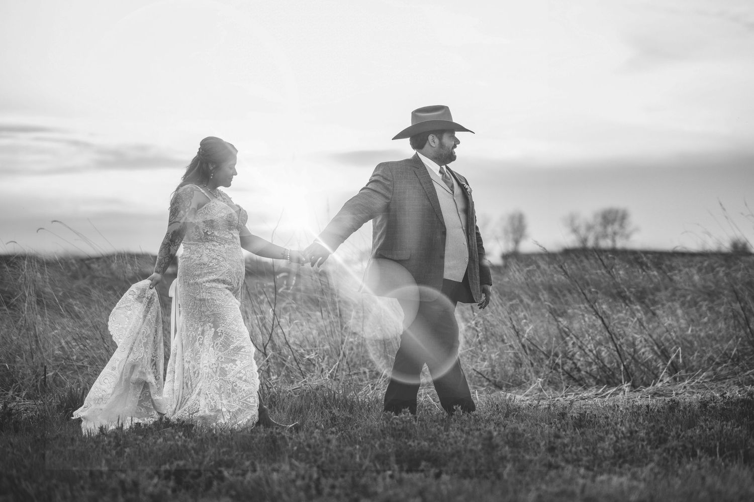Two people in wedding attire walking together through a rustic field at dusk in black and white photography.