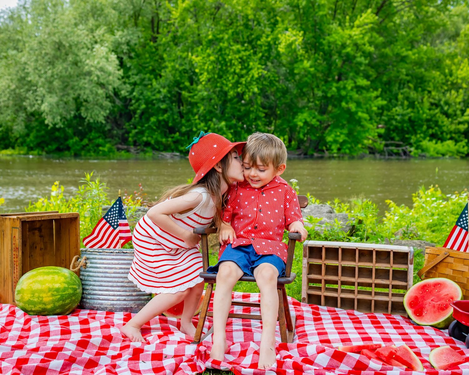 Children in patriotic outfits share a picnic with watermelon and American flags by a lake.