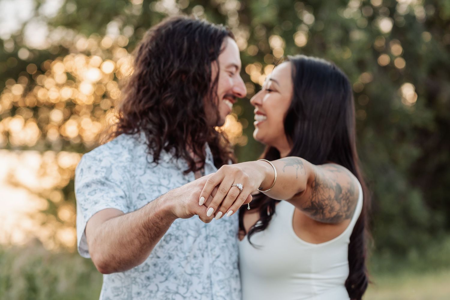 Happy couple sharing an intimate moment while showing engagement ring in a sunlit outdoor setting at sunset.