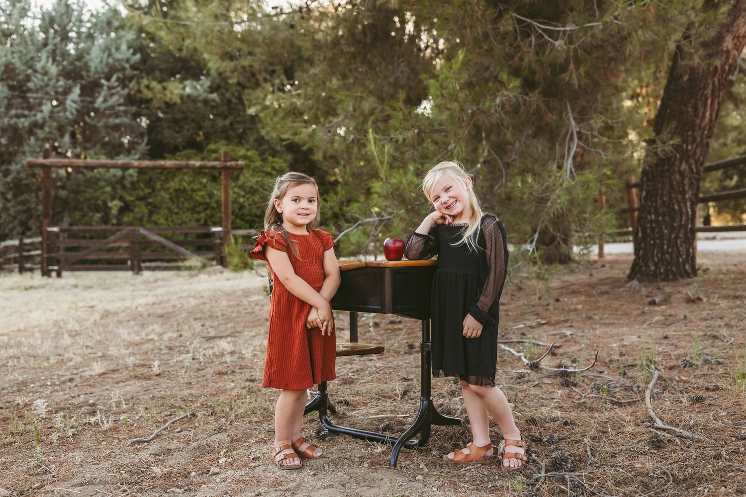Two young children in dresses stand by a black metal music stand on natural outdoor ground.