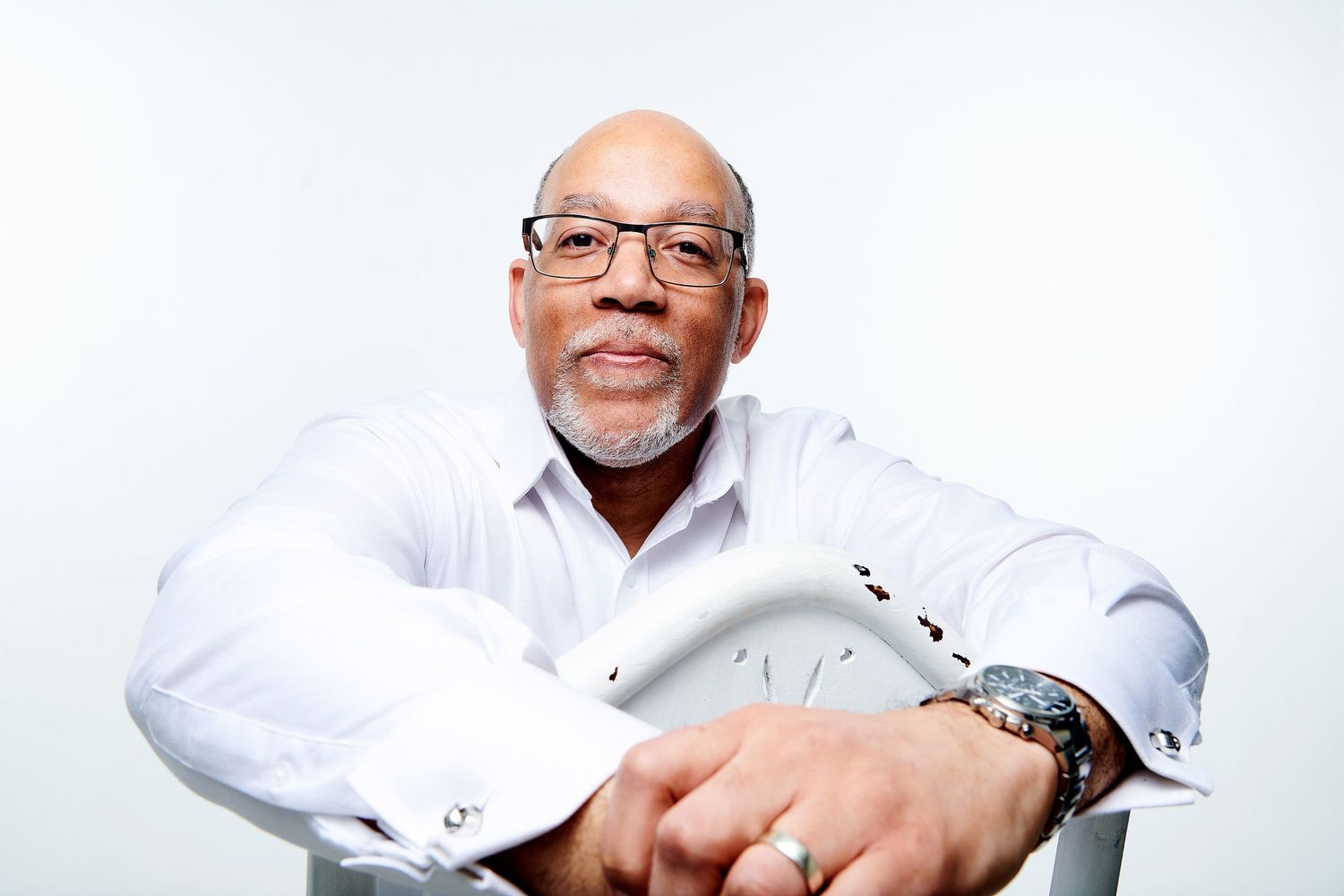Professional senior in glasses wearing white button down shirt sitting with arms crossed against white background.