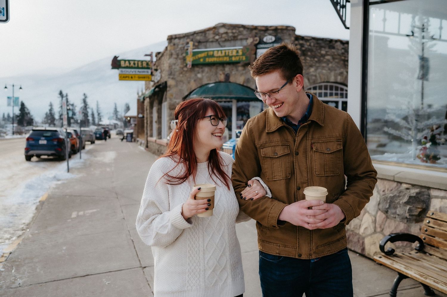 Winter street scene with bundled up couple looking at phone outside rustic buildings.