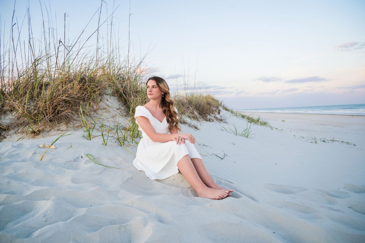 Person in white dress sits peacefully on sandy beach dunes at sunset with sea grass and ocean in background. Myrtle Beach Photographer