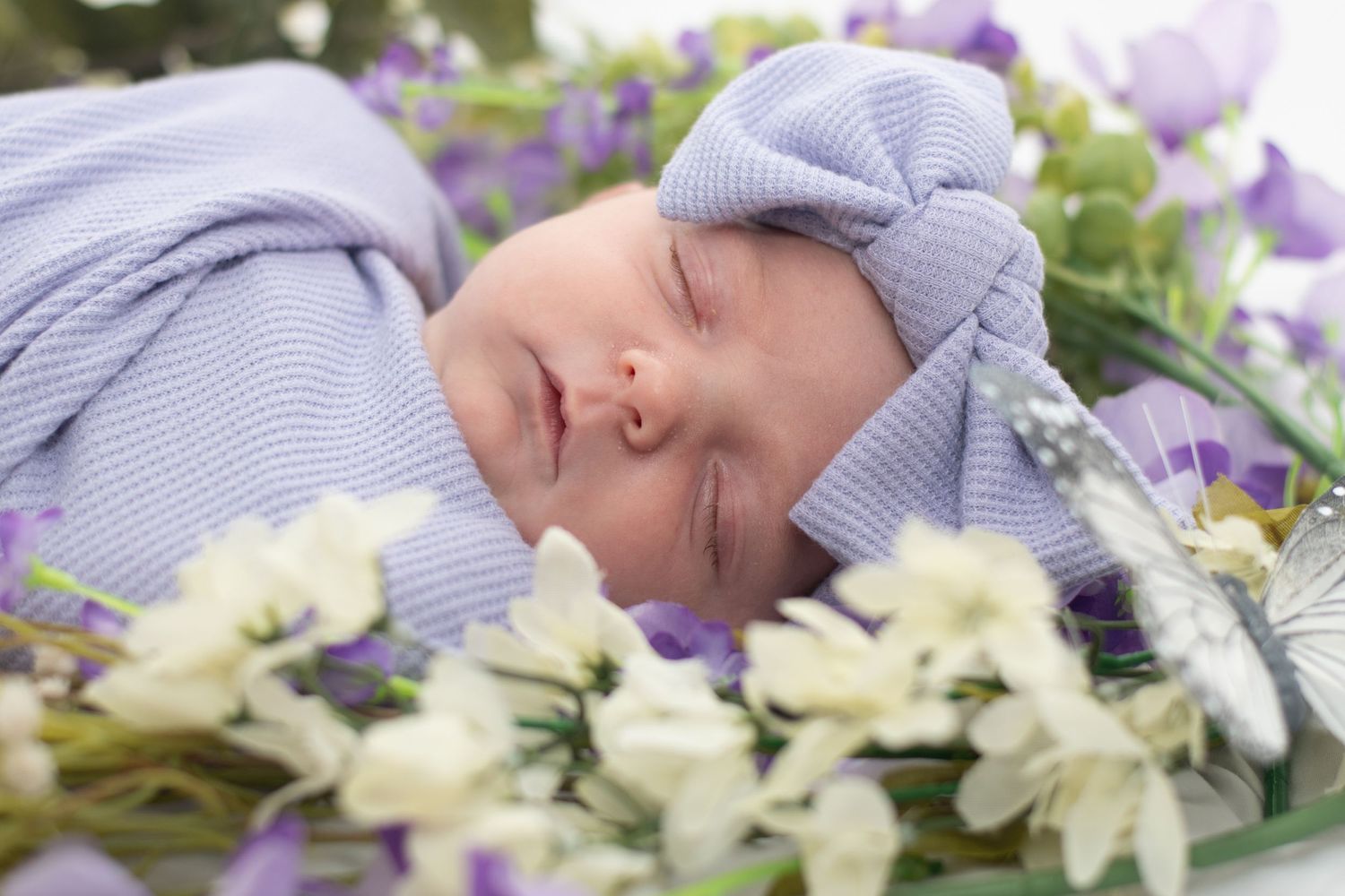 Sleeping baby wearing purple hat nestled among spring flowers and blossoms.