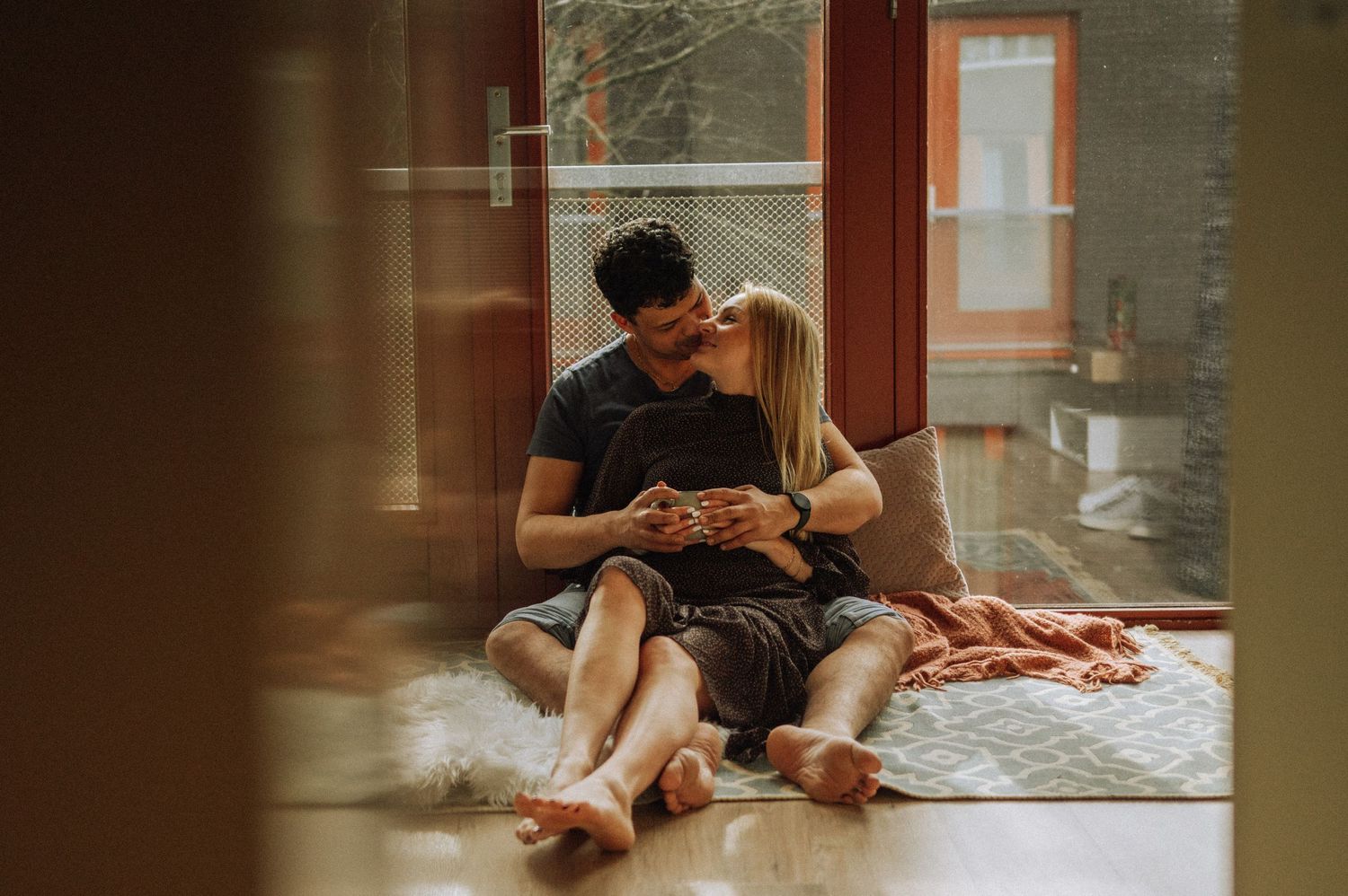 A couple sits intimately on the floor by a window sharing a tender moment in warm sunlight.