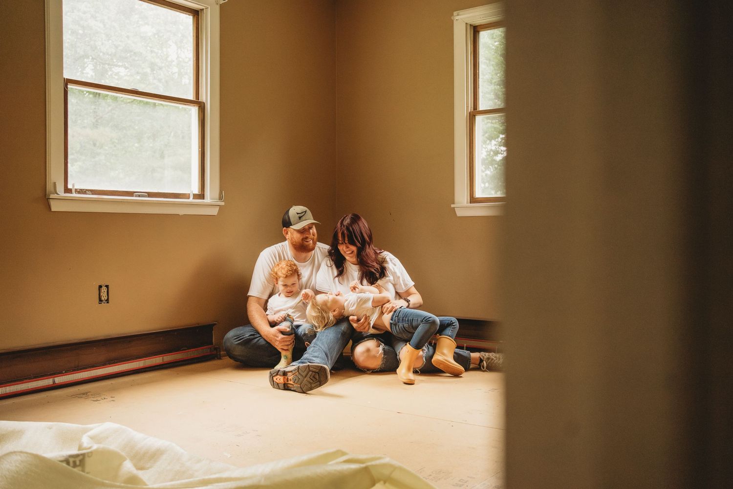 Family sits together on floor of empty room with sunlight streaming through windows.