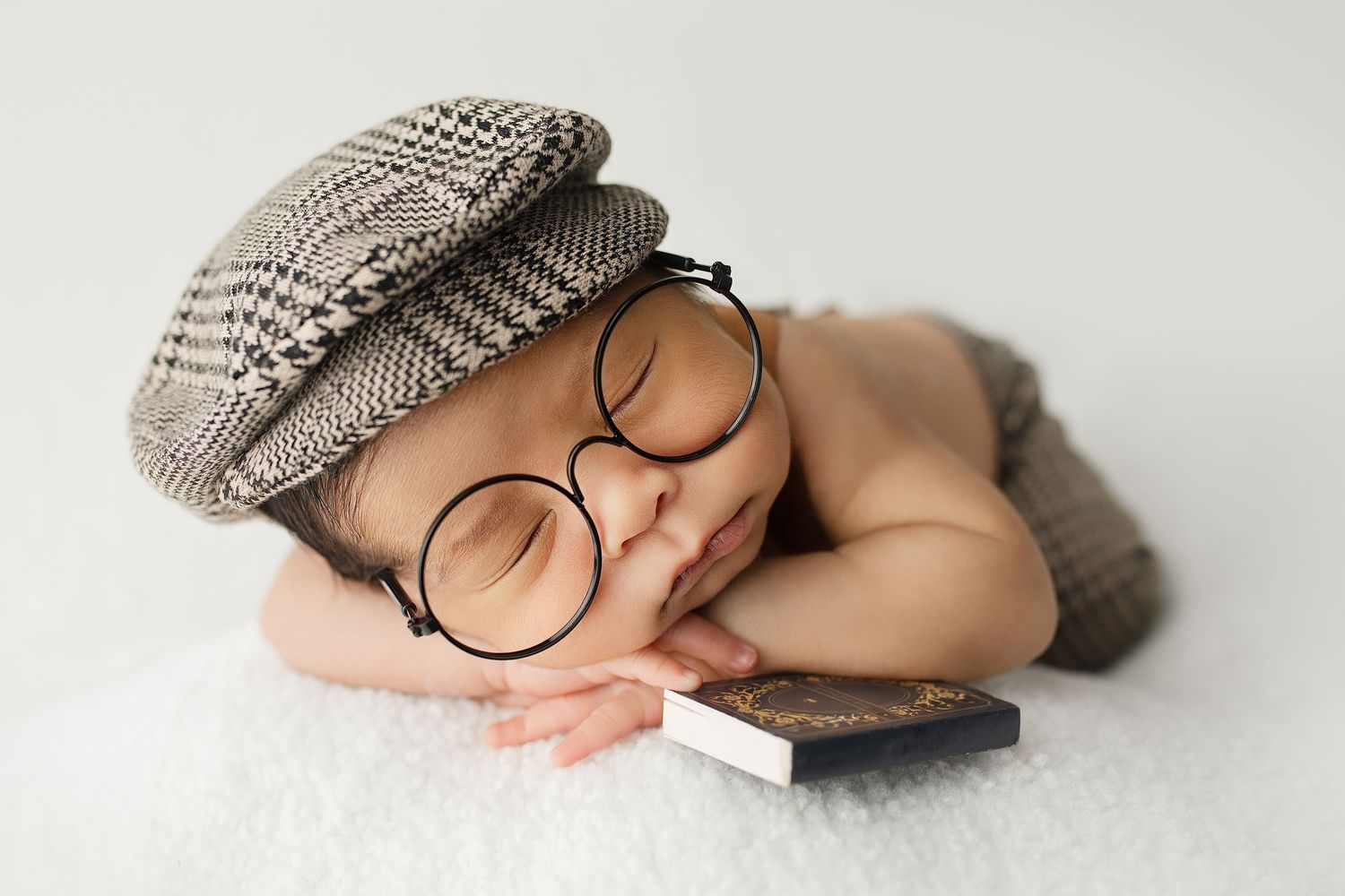 A sleeping newborn wearing round glasses and a newsboy cap rests peacefully on a white surface next to a book.
