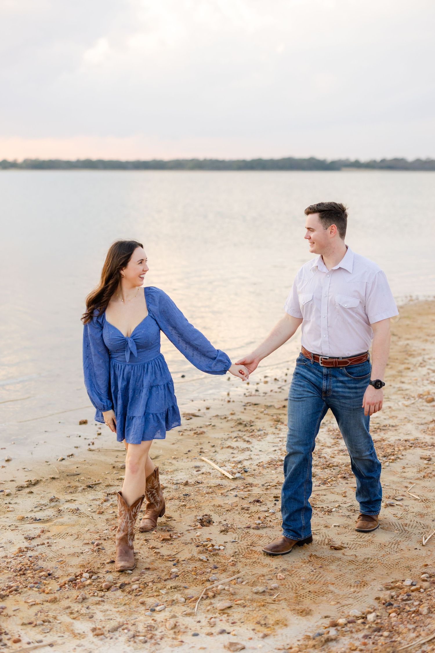 Couple in casual western attire holding hands while walking along a serene beach at sunset.
