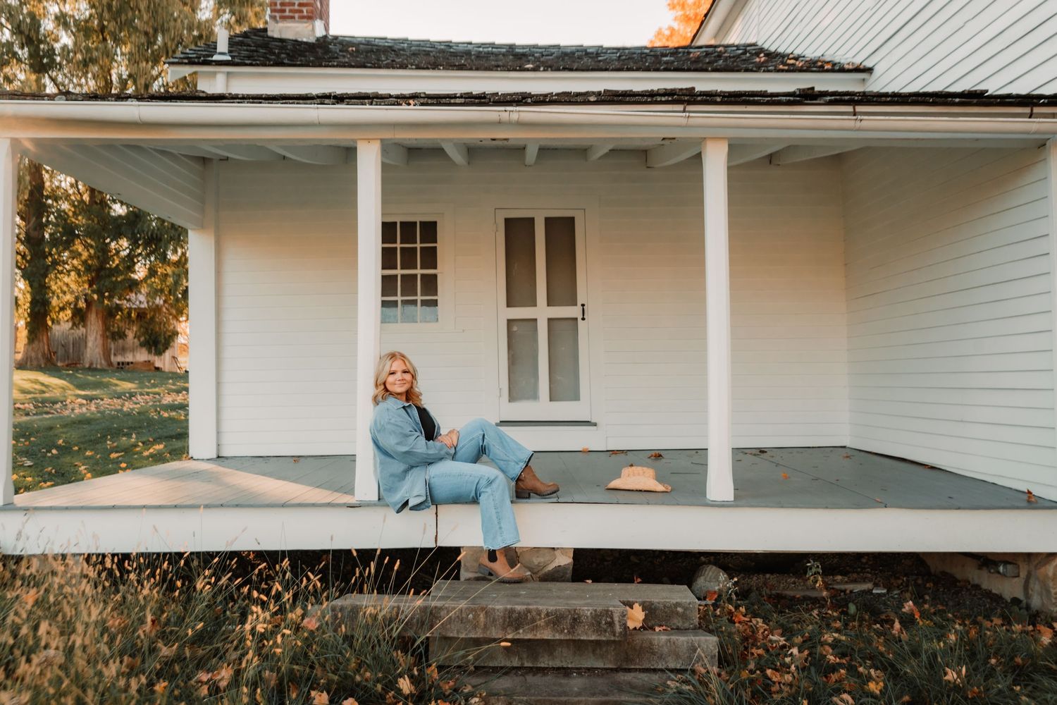 A person sits relaxed on the porch of a white farmhouse with pillars at sunset.