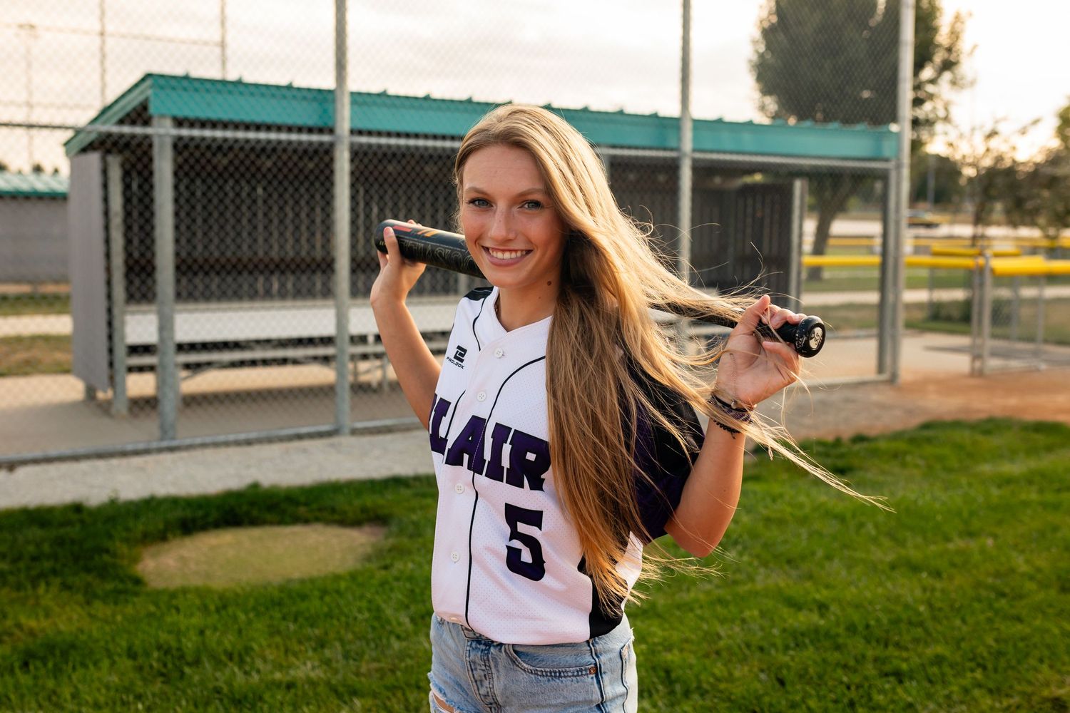 Softball player in white uniform poses with bat on field at sunset.