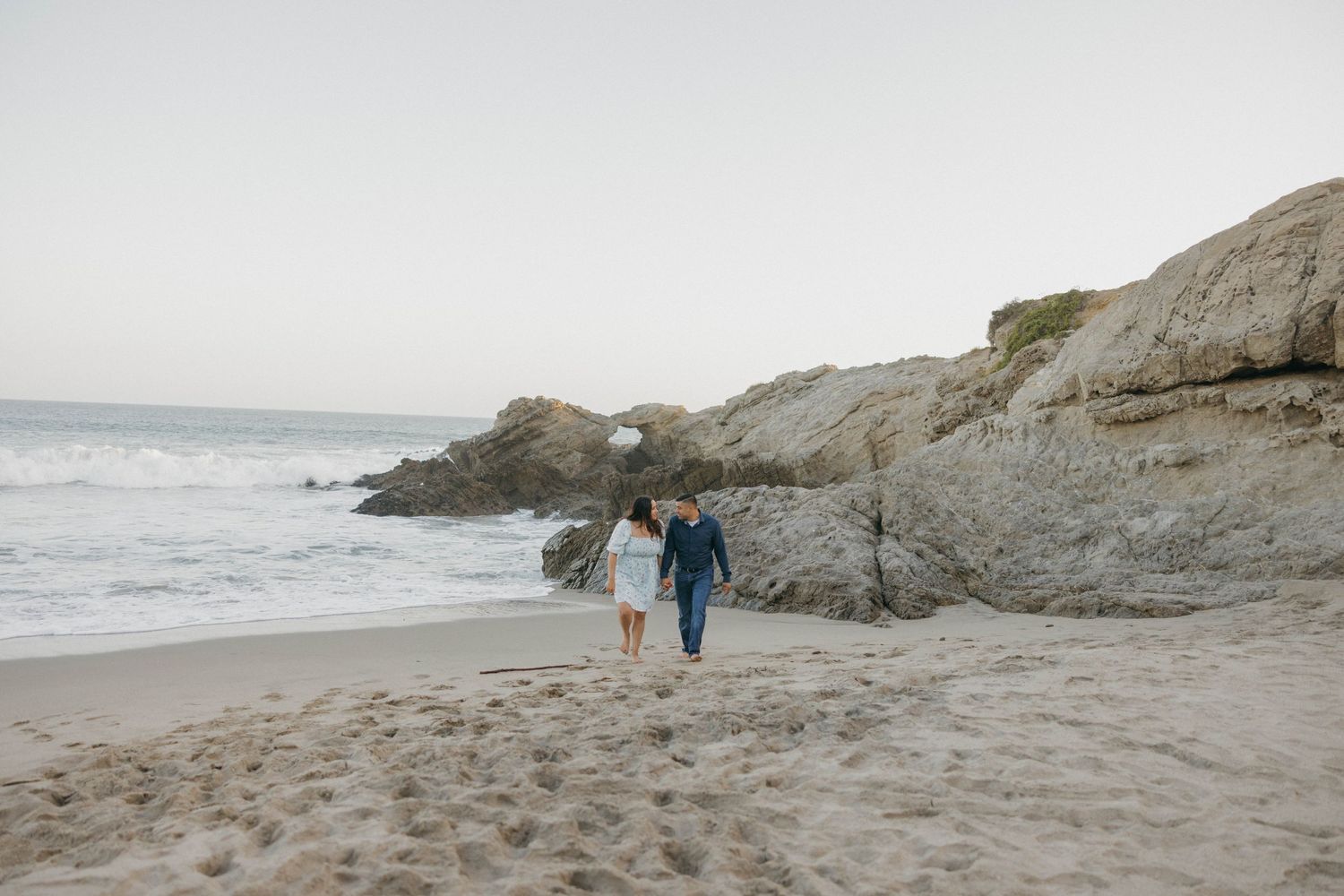 A couple walking hand in hand along a rocky beach with dramatic coastal cliffs in the background.