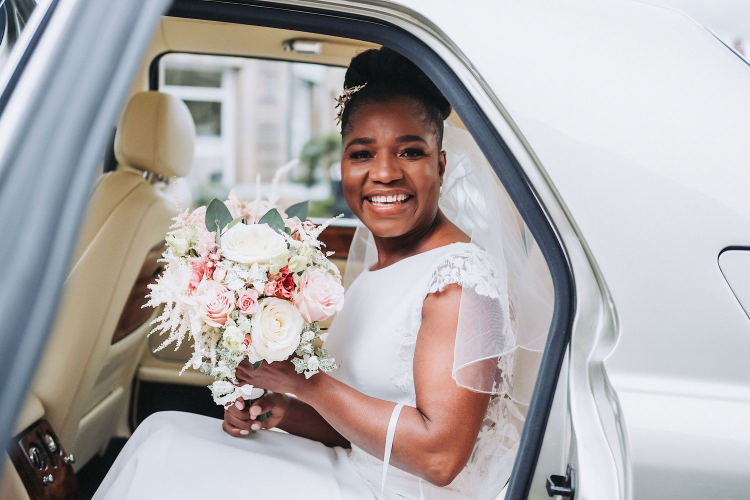 A joyful bride in white holding a pink and white bouquet while seated in a car.