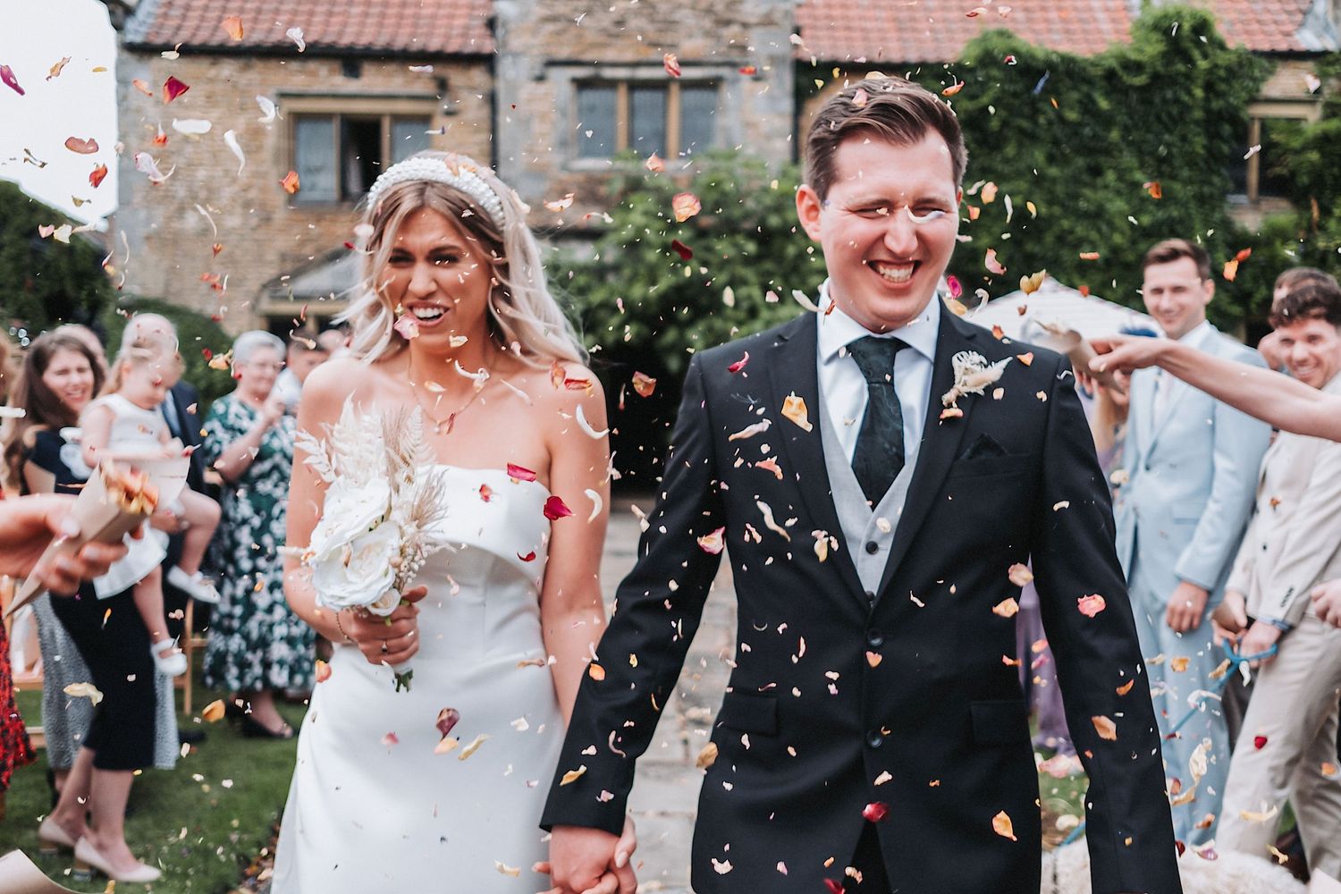 Happy newlyweds walking through confetti shower at rustic wedding venue with guests in background.