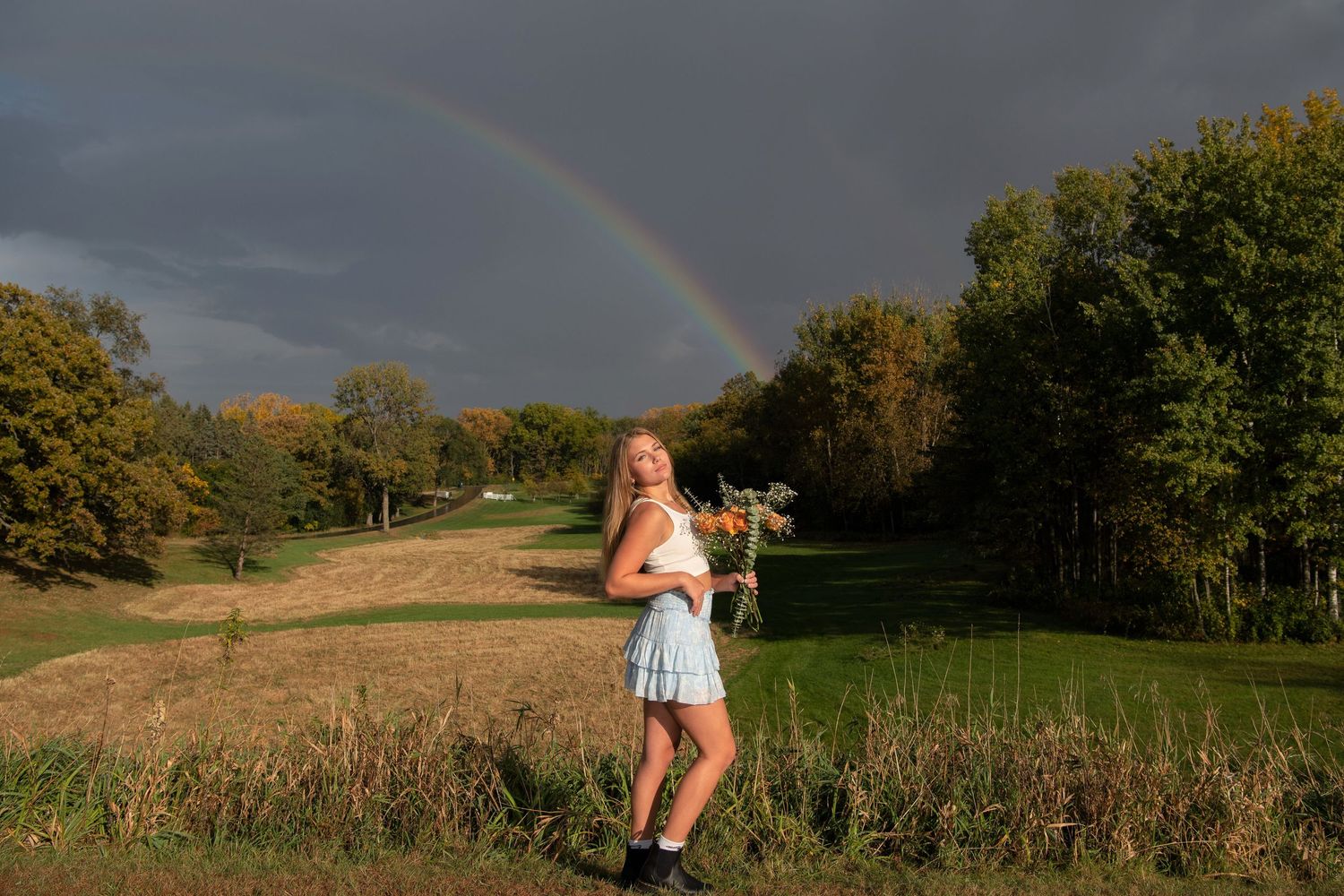 A person stands in a field under a colorful rainbow arch while holding flowers on a late summer afternoon.