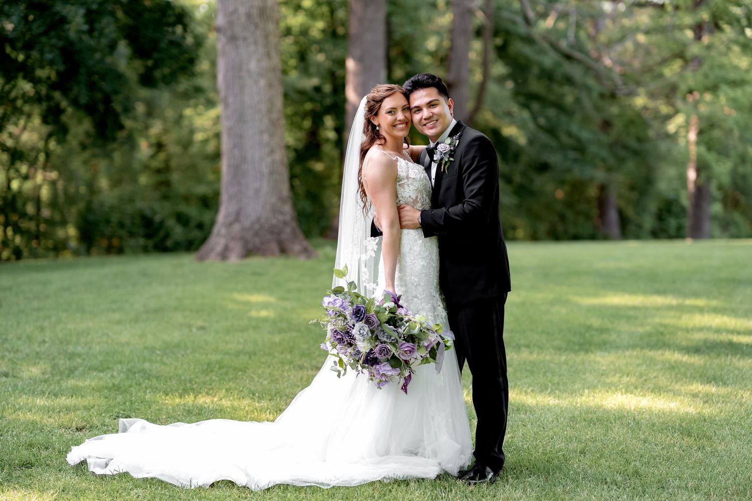 Bride and groom embracing outdoors on green lawn, bride holding purple bouquet in white gown, groom in black tuxedo.