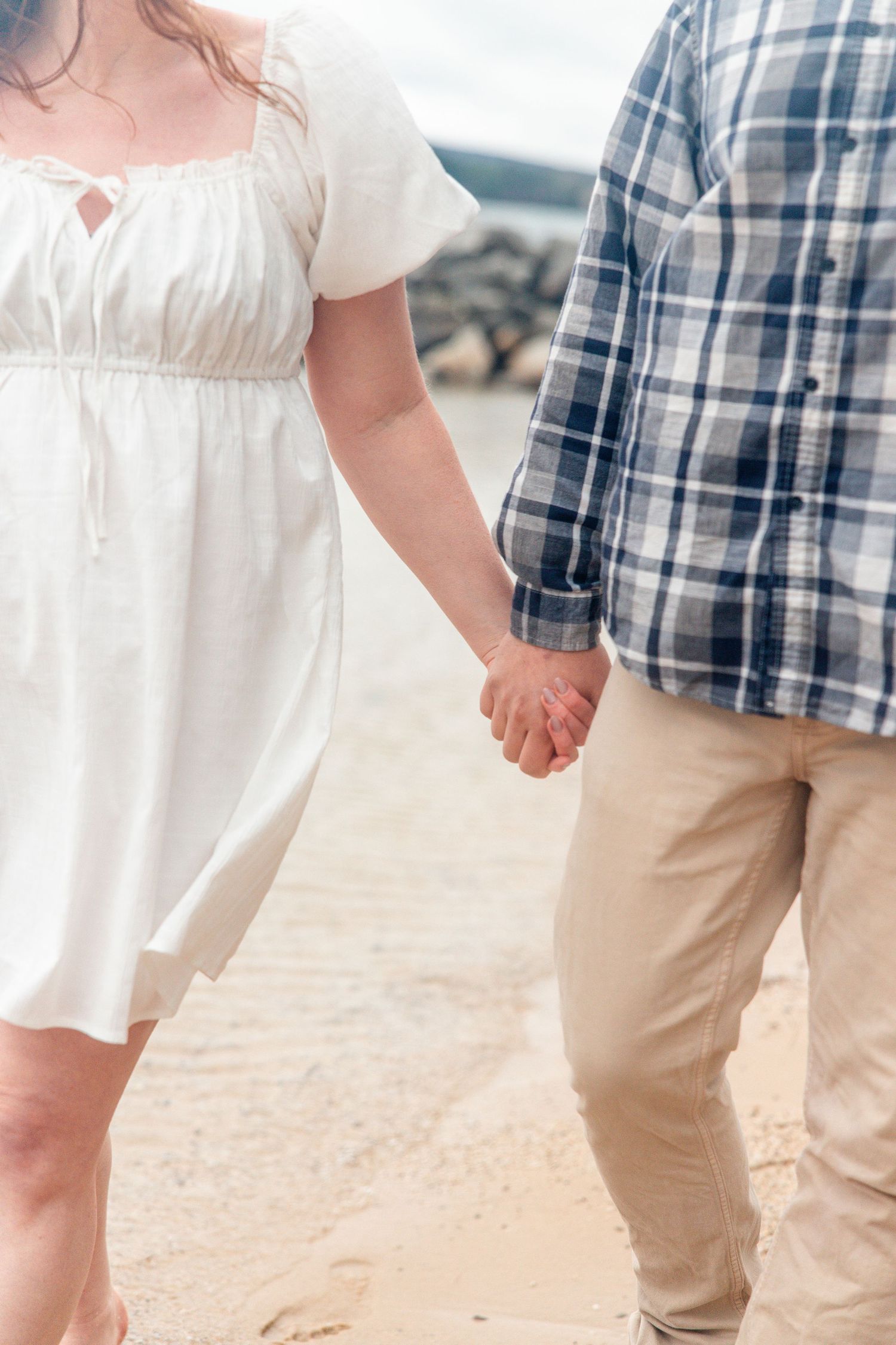 Couple holding hands on a sandy beach, woman in white dress and man in plaid shirt with khaki pants.