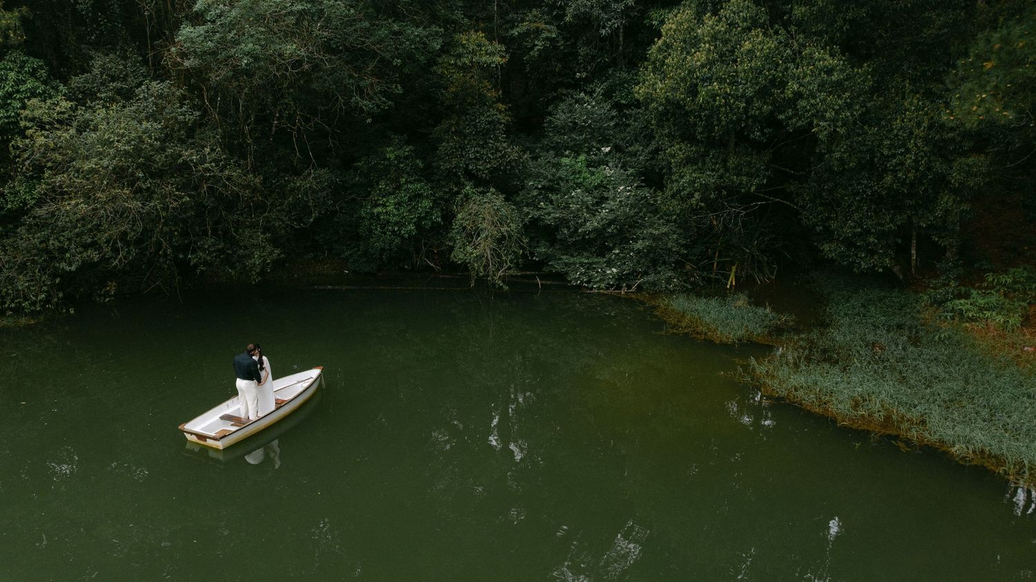 Aerial view of a couple standing on a white boat surrounded by lush green forest on a calm green lake.