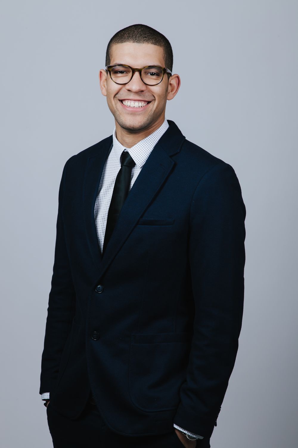 Young professional man in dark suit, tie, and glasses smiling confidently against gray background.