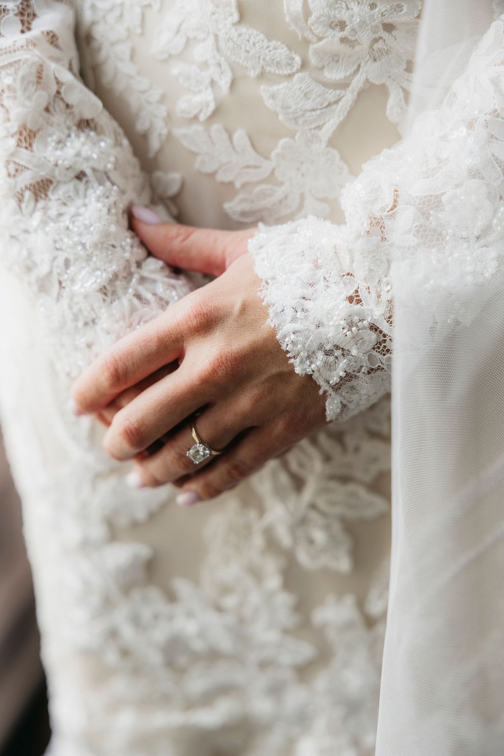 Bride wearing a diamond engagement ring on her hand, dressed in an elegant white lace long-sleeve wedding gown.