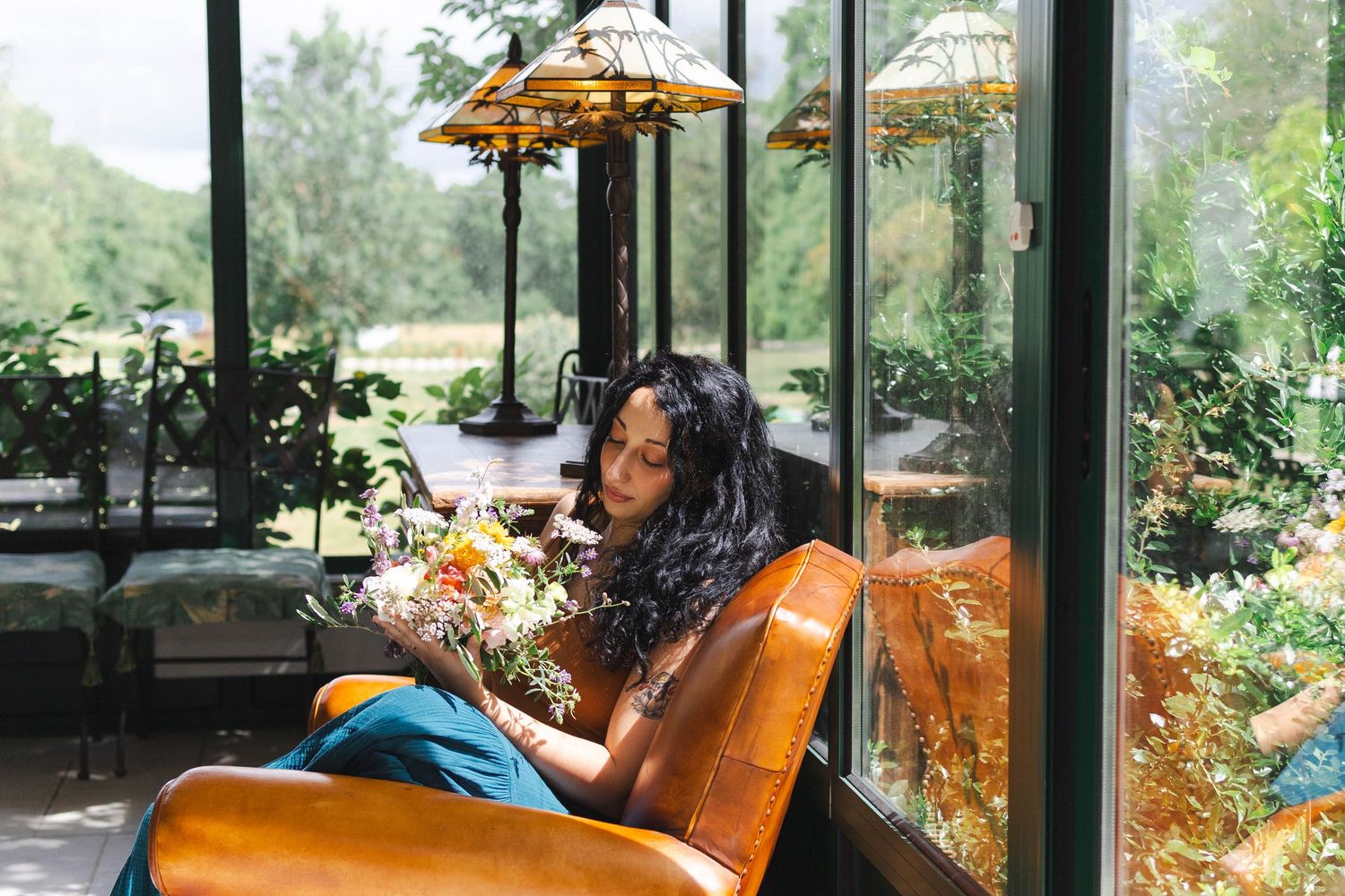 Woman sitting in leather chair holding wildflowers in a sunlit greenhouse with lush greenery and stained glass lamps.