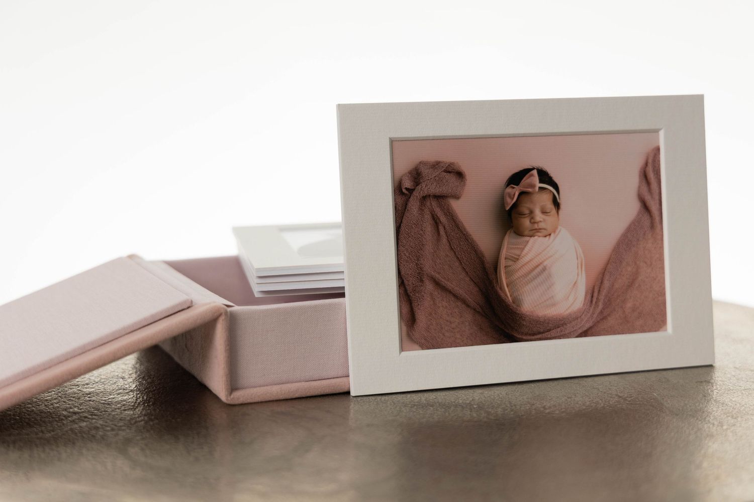 Pink keepsake box with framed newborn photo of swaddled baby girl with floral headband on gold surface.