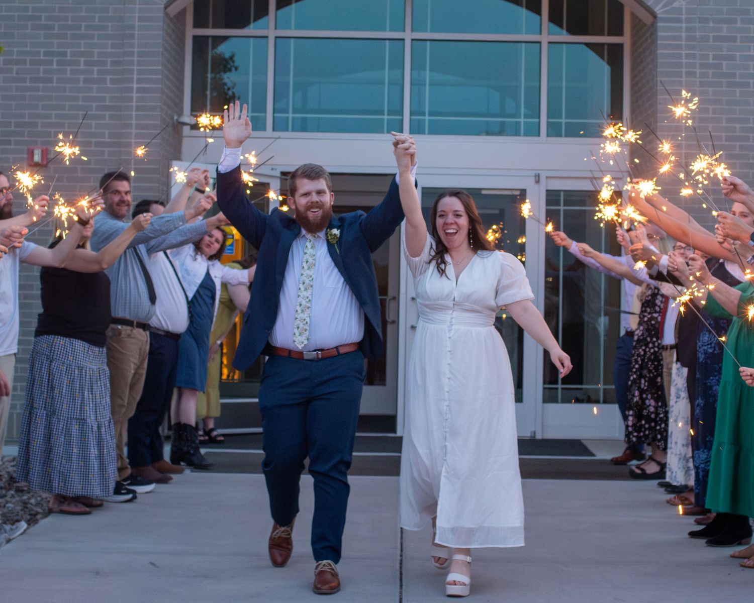 Wedding guests create a magical sparkler exit tunnel at dusk outside a brick building during the celebration.