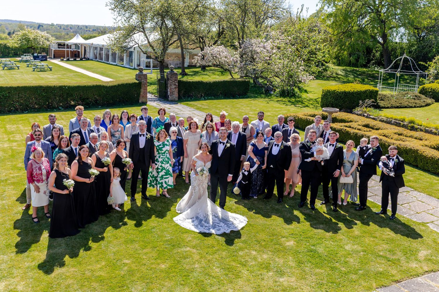 Large wedding group photo outdoors on sunny day, bride and groom centered among guests on manicured lawn with gardens.