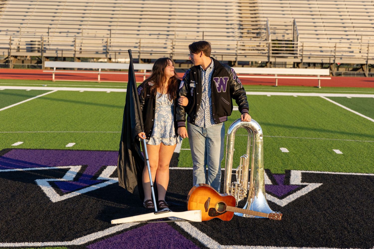 Two high school band members pose on a football field with a flag, trombone, and guitar in purple school colors.