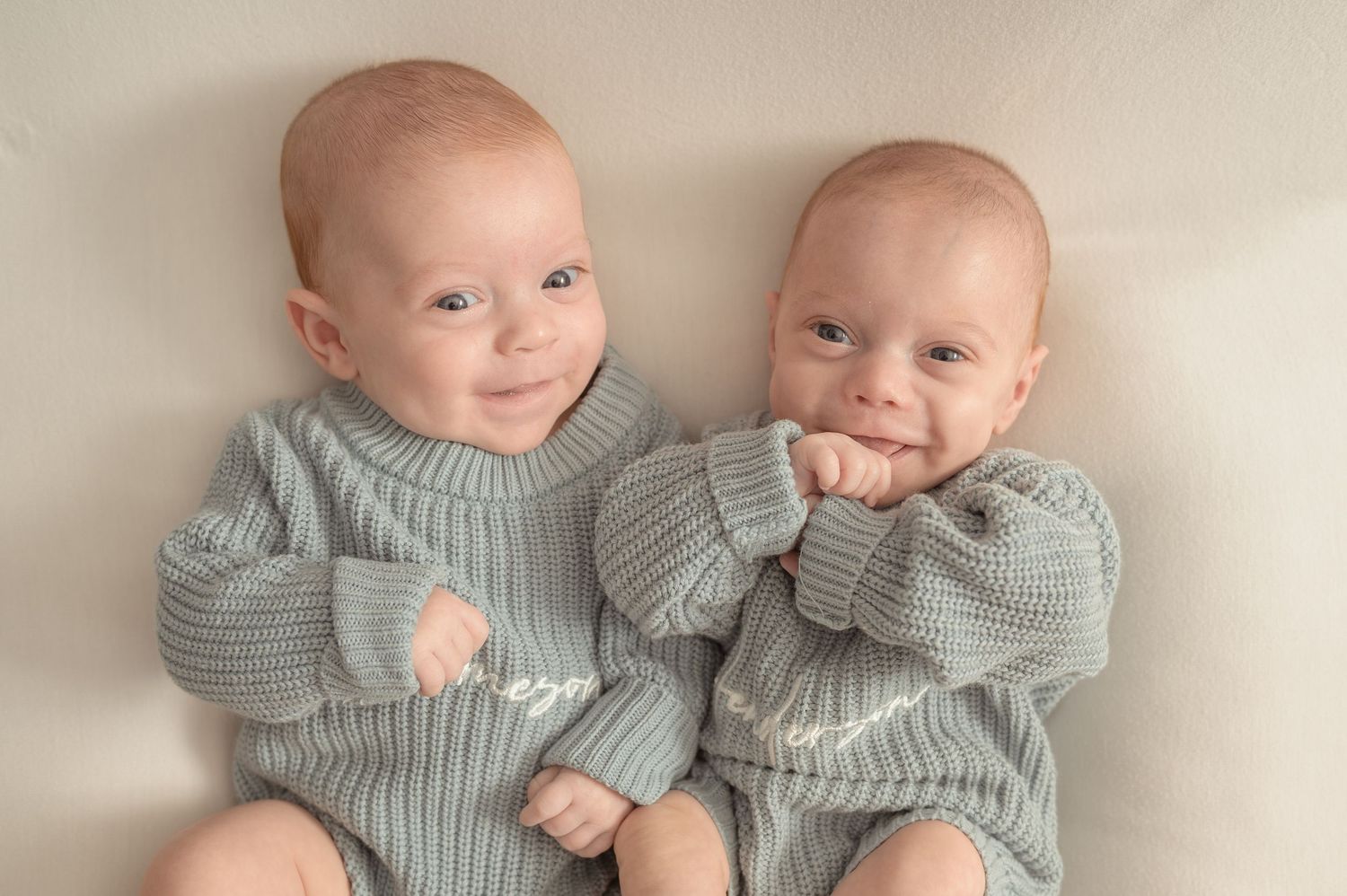A young couple in white shirts and jeans sit on a play mat smiling at their two babies lying on their stomachs.